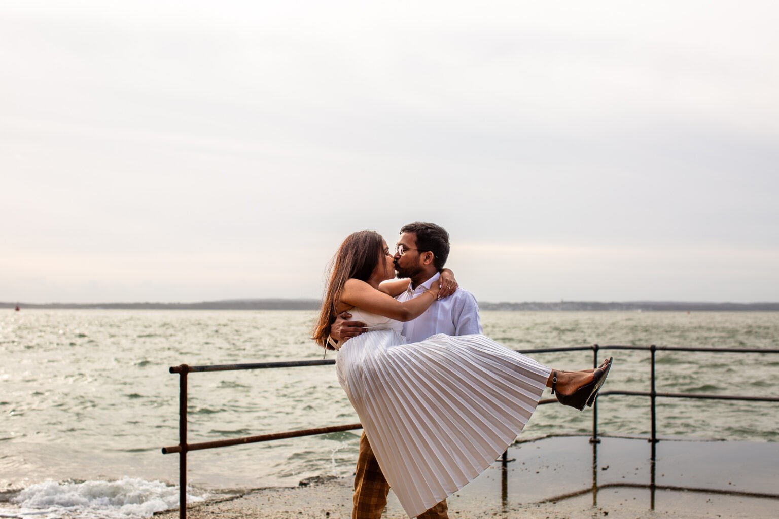 Groom is holding the bride at his arms and is kissing her at the beach in Old Portsmouth during couple photography.