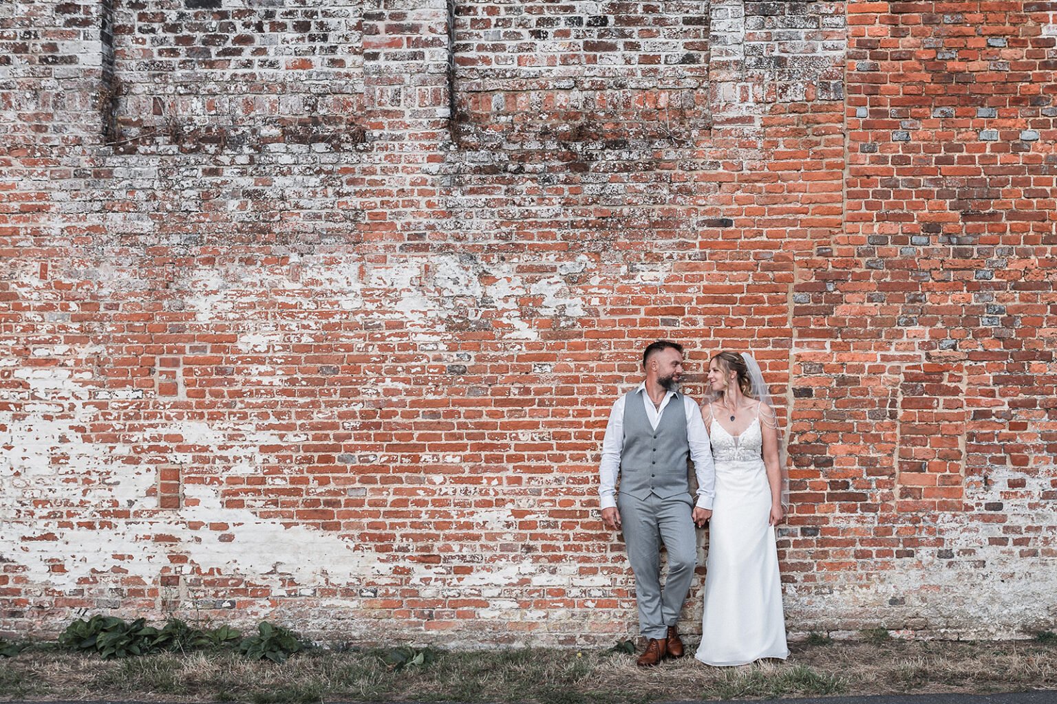 Bride and groom are looking at each other in front of a brick wall outside Cross Barn in Odiham during wedding photography.